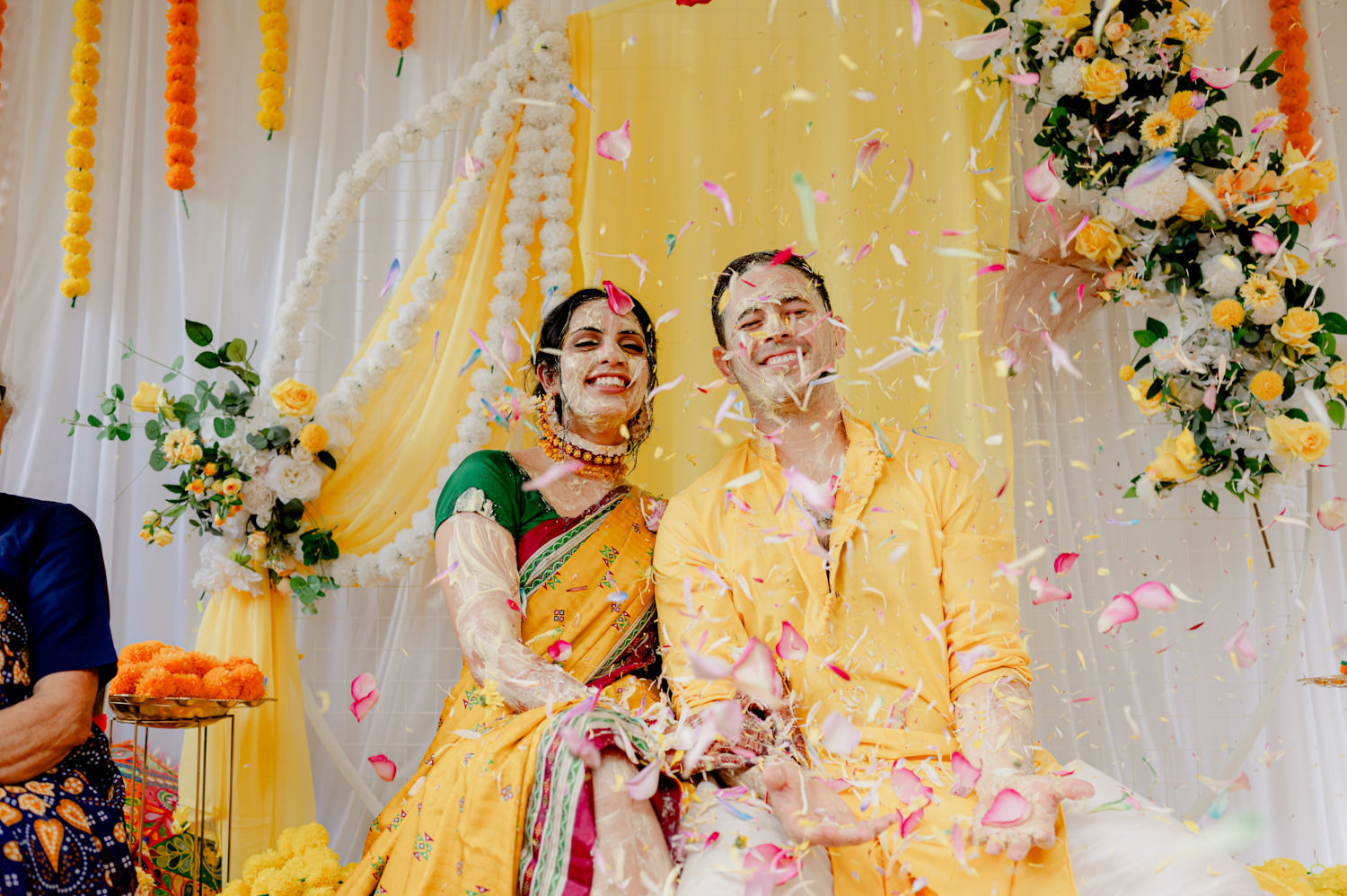 A couple in colourful Indian clothes smile as flower petals are thrown in the air