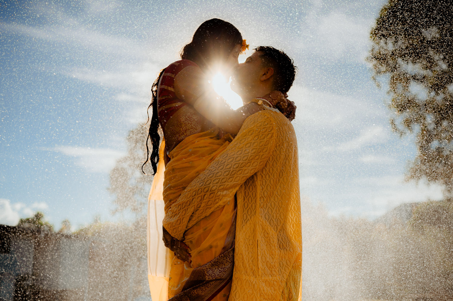A couple wearing yellow Indian clothing kiss underneath a shower of water with the sun behind them